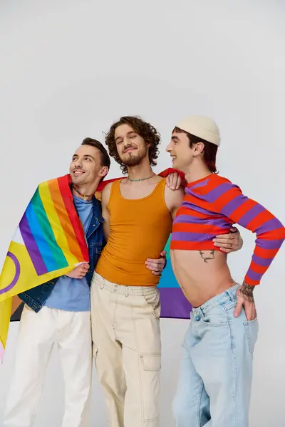 Three appealing cheerful gay men in cozy clothing posing actively with rainbow flag on gray backdrop — Stock Photo