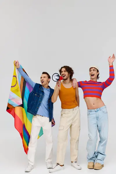 Three cool joyous gay men in cozy outfit holding rainbow flag and using megaphone on gray backdrop — Stock Photo