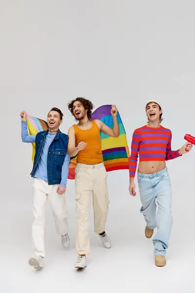 Three trendy joyous gay men in cozy outfit holding rainbow flag and using megaphone on gray backdrop — Stock Photo