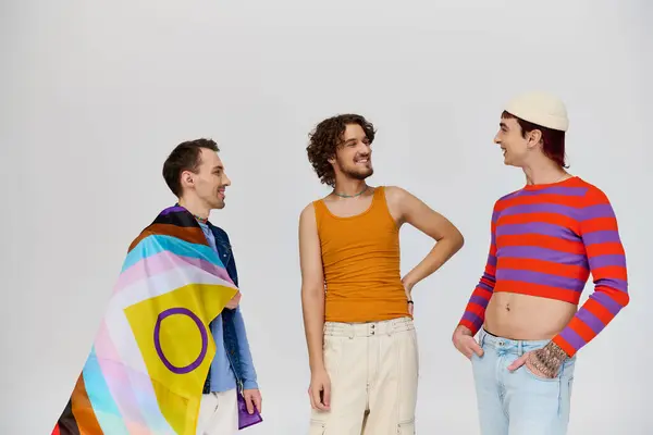 Drei gut aussehende, fröhliche schwule Männer in kuscheliger Kleidung posieren aktiv mit Regenbogenfahne vor grauem Hintergrund — Stockfoto