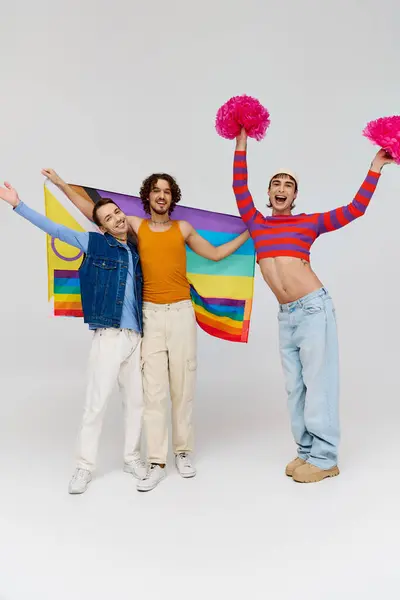 Позитивные привлекательные геи в яркой одежде позируют с радужным флагом и помпонами на сером фоне — стоковое фото