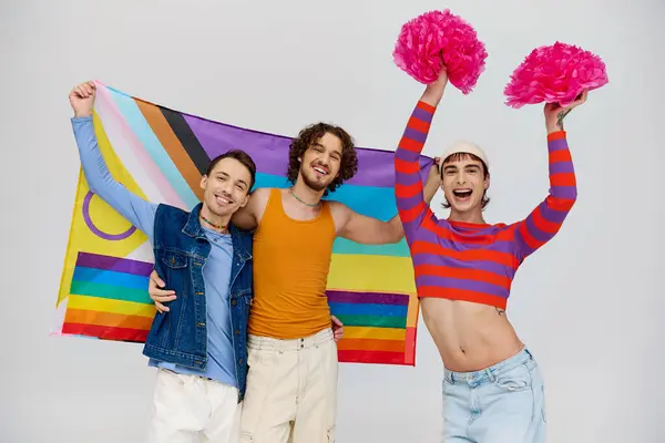Allegro attraente gay gli uomini in vibrante vestiti posa con arcobaleno bandiera e pon pon su grigio sfondo — Foto stock