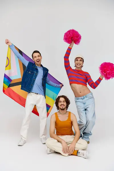 Весело привлекательные геи в яркой одежде позируют с радужным флагом и помпонами на сером фоне — стоковое фото