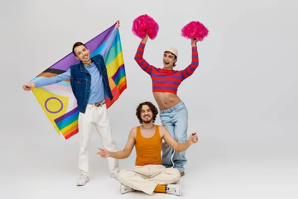 Joyful appealing gay men in vibrant clothes posing with rainbow flag and pom poms on gray backdrop — Stock Photo