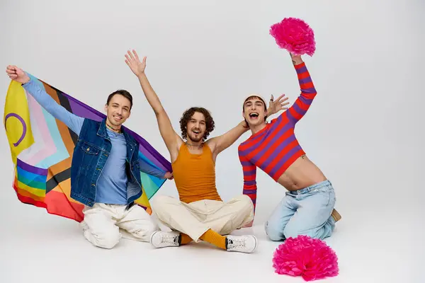 Радостные привлекательные геи в яркой одежде, позирующие с радужным флагом и помпонами на сером фоне — стоковое фото