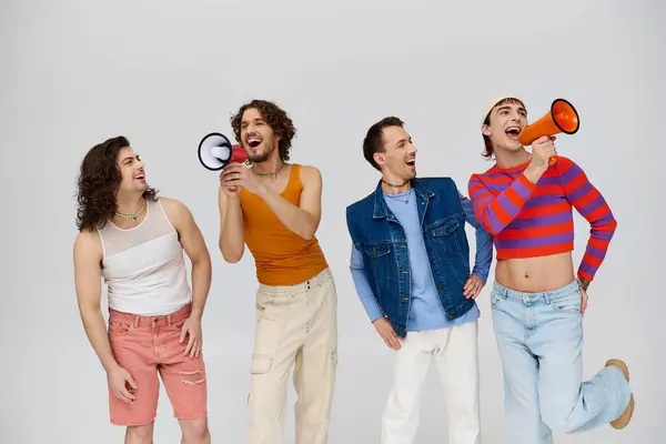 Four cheerful fancy gay men in stylish outfits using megaphones and posing actively on gray backdrop — Stock Photo