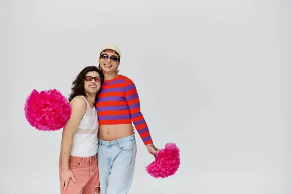 Appealing jolly gay men in vibrant attires with sunglasses posing with pom poms on gray backdrop — Stock Photo