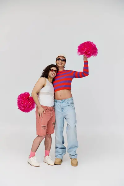 Guapo alegre gay los hombres en vibrante atuendos con gafas de sol posando con pompones en gris telón de fondo - foto de stock