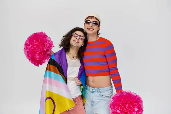 Dos alegre seductor gay los hombres en negrita trajes con gafas de sol posando con arco iris bandera y pompones - foto de stock