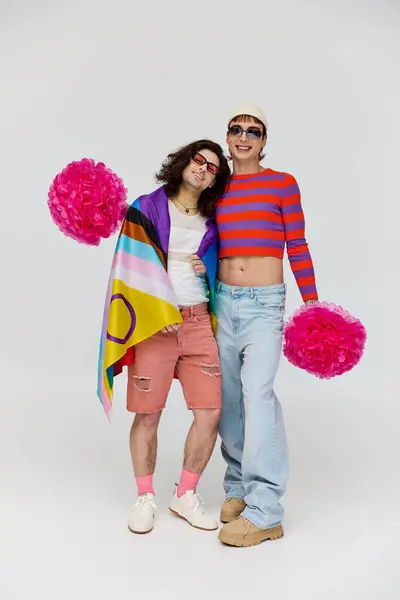 Two joyous alluring gay men in bold attires with sunglasses posing with rainbow flag and pom poms — Stock Photo