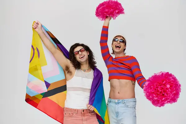Дві радісні привабливі геї в сміливих вбраннях з сонцезахисними окулярами позує з веселковим прапором і помпою — стокове фото