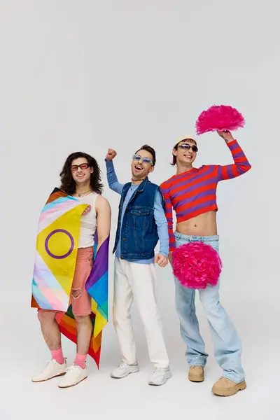 Three jolly stylish gay men in bold clothes with sunglasses posing with pom poms and rainbow flag — Stock Photo