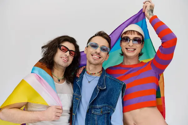 Positif attrayant gay les hommes avec des lunettes de soleil à la mode tenant drapeau arc-en-ciel et souriant à la caméra — Photo de stock