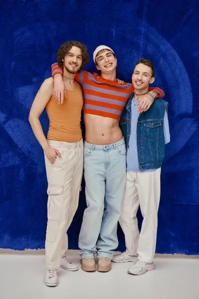 Three stylish appealing merry gay friends in cozy attires posing on dark blue backdrop, pride month — Stock Photo