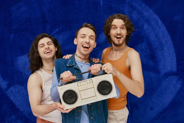 Tres jóvenes alegre gay amigos posando con cinta grabadora en azul oscuro telón de fondo, orgullo mes - foto de stock