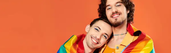 Jolly appealing gay men in cozy attires with rainbow flag on orange backdrop, pride month, banner — Stock Photo