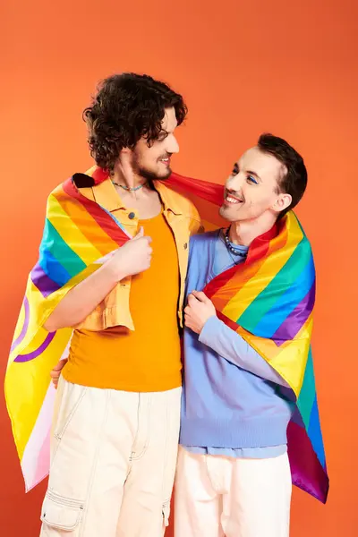 Joyous appealing young gay men in cozy attires with rainbow flag on orange backdrop, pride month — Stock Photo