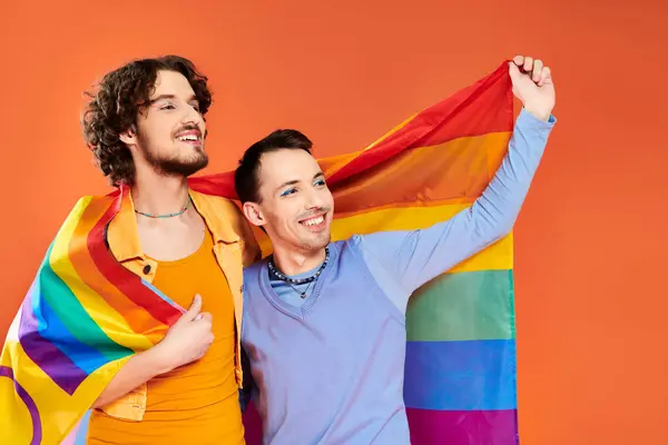 Cheerful appealing young gay men in cozy attires with rainbow flag on orange backdrop, pride month — Stock Photo