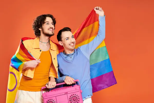 Two joyful handsome gay friends posing with tape recorder and rainbow flag on orange backdrop — Stock Photo