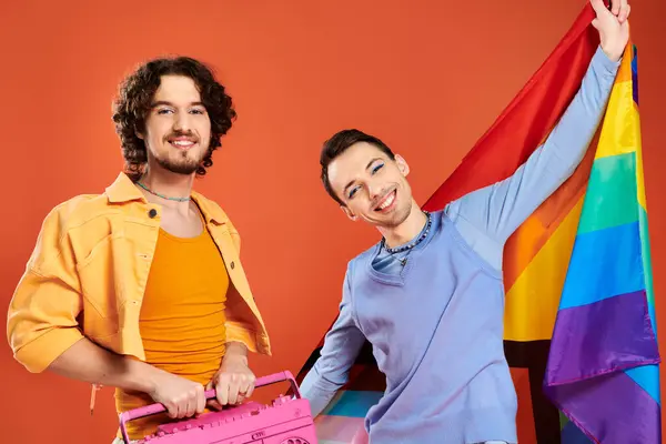 Dos contento guapo gay amigos posando con cinta grabadora y arco iris bandera en naranja telón de fondo - foto de stock