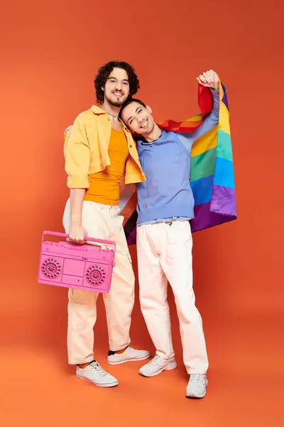 Dos positivo guapo gay amigos posando con cinta grabadora y arco iris bandera en naranja telón de fondo - foto de stock