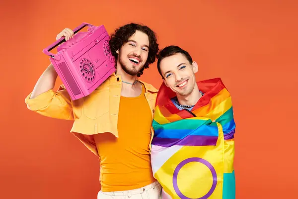 Dos alegre guapo gay amigos posando con cinta grabadora y arco iris bandera en naranja telón de fondo - foto de stock