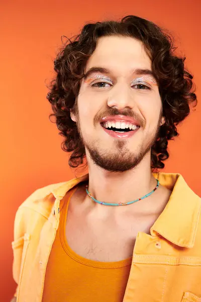 Alegre atractivo gay hombre con oscuro cabello y vibrante maquillaje posando en naranja telón de fondo, orgullo mes - foto de stock