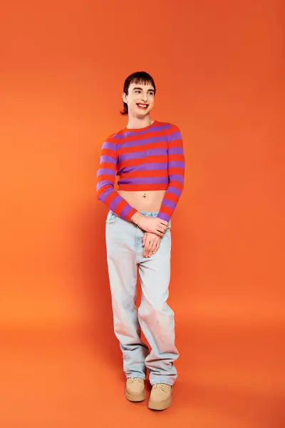 Jolly appealing gay man in vibrant clothes with makeup posing on orange backdrop and looking away — Stock Photo