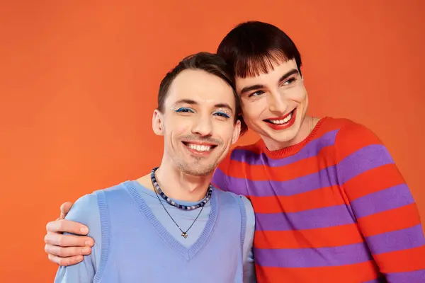 Alegre atractivo gay amigos con vívido maquillaje posando juntos en naranja telón de fondo, orgullo mes - foto de stock