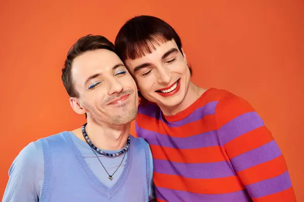 Positive attractive gay friends with vivid makeup posing together on orange backdrop, pride month — Stock Photo
