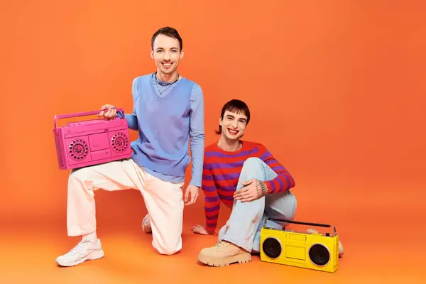 Deux joyeux beau gay les hommes avec vibrant maquillage posant avec des magnétophones sur fond orange — Photo de stock