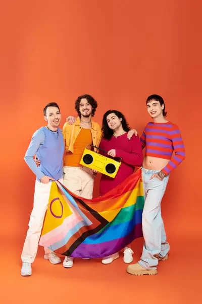 Four young merry gay friends posing with rainbow flag and tape recorder on orange background — Stock Photo