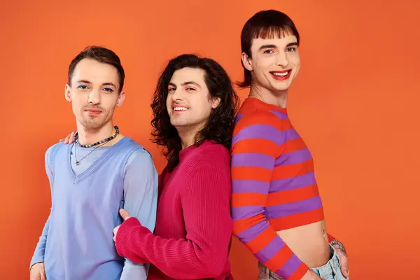 Tres amigos gays alegres sofisticados en ropa vívida posando juntos sobre fondo naranja, orgullo - foto de stock