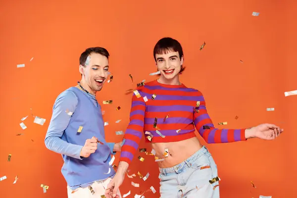 Alegre atractivo gay los hombres en vibrantes atuendos con maquillaje posando activamente en naranja telón de fondo - foto de stock