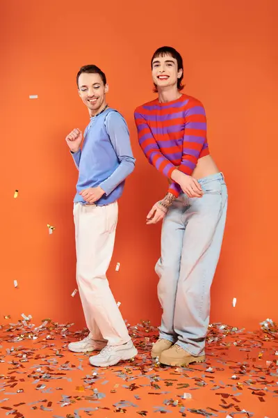 Zwei fröhliche, stylische schwule Männer in lebendigen Anzügen, die sich unter Konfettiregen auf orangefarbenem Hintergrund amüsieren — Stockfoto