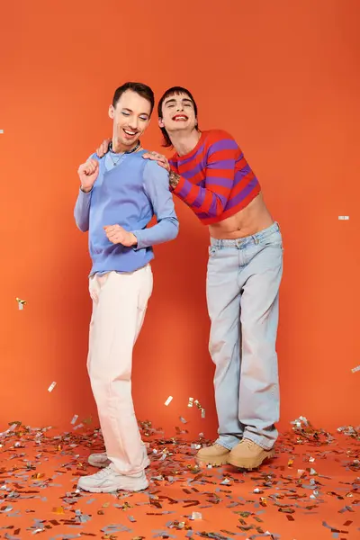 Deux gays élégants joyeux en tenue vibrante s'amusant sous la pluie de confettis sur fond orange — Photo de stock