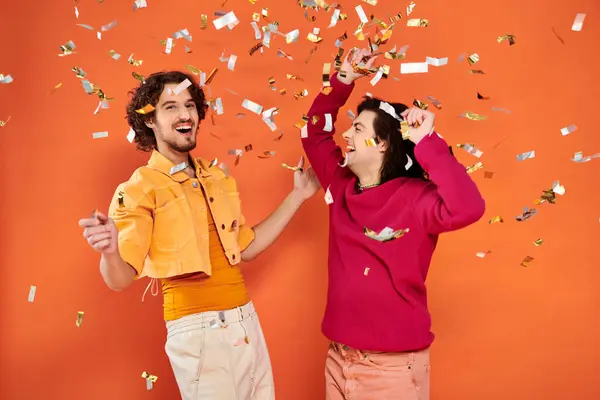 Dos alegre elegante gay los hombres en vibrante atuendos tener divertido bajo confeti lluvia en naranja fondo - foto de stock