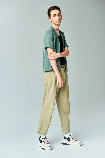 Ein junger queerer Mensch posiert selbstbewusst in einem Studio, bekleidet mit grünem Hemd und khakifarbener Hose, vor grauem Hintergrund. — Stockfoto