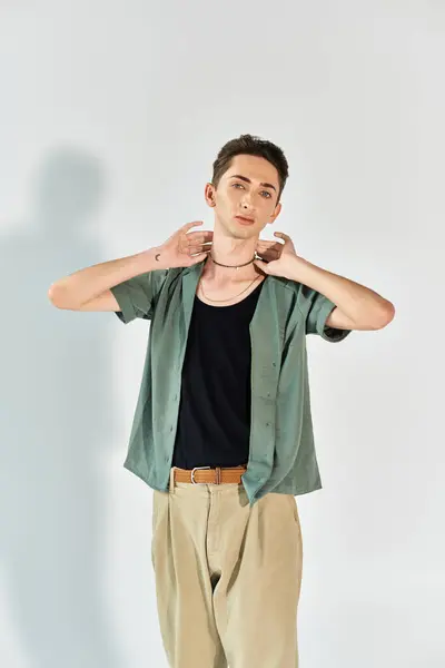 Una giovane persona queer in uno studio, indossa una camicia verde e pantaloni abbronzanti, trasudando orgoglio e fiducia in uno sfondo grigio. — Foto stock