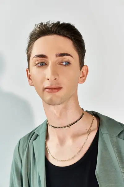 Eine junge queere Person posiert selbstbewusst in einer stylischen grünen Jacke und einer auffälligen Halskette vor grauem Studiohintergrund. — Stockfoto