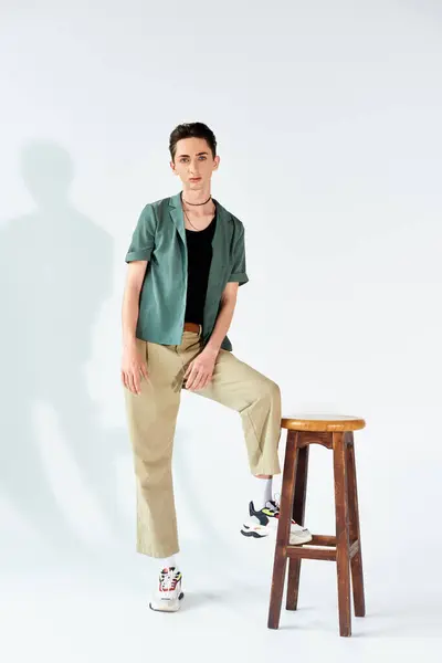 Una joven queer con estilo se para con confianza en un taburete con una camisa verde y pantalones caqui en un estudio sobre un fondo gris. — Stock Photo