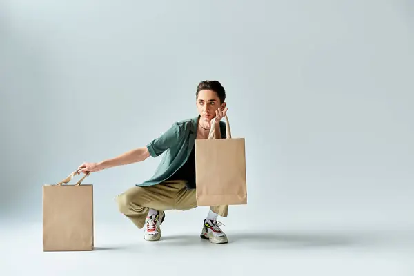 A young queer crouches, exuding pride, holding shopping bags in a studio against a grey background. — Stock Photo