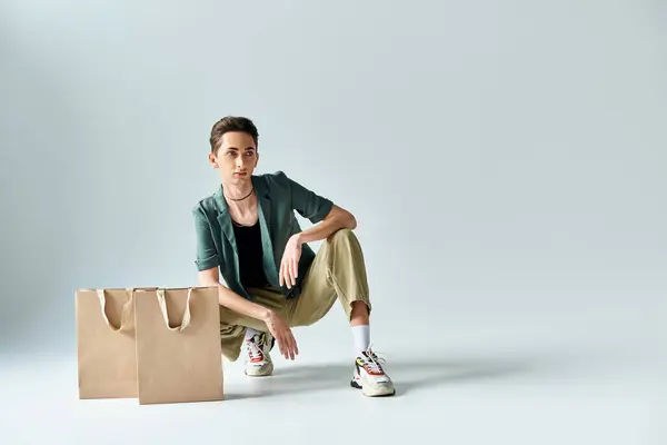 A young queer person sitting on the floor surrounded by colorful shopping bags in a studio on a grey background. — Stock Photo