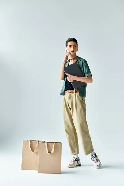 Стильный молодой человек без особых усилий балансирует сумки и мобильный телефон, источая уверенность и городской стиль на сером фоне. — стоковое фото
