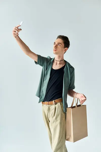 Un joven queer sostiene una bolsa de compras, capturando una selfie en un estudio sobre un fondo gris. — Stock Photo
