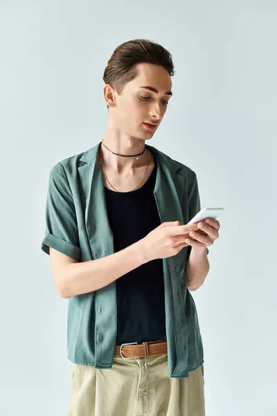 A stylish young queer man, wearing a green shirt and tan pants, checks his phone against a grey studio backdrop. — Stock Photo