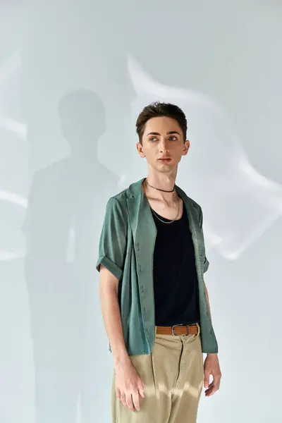 Um jovem queer faz uma pose confiante em uma camisa verde elegante e calças bronzeadas contra uma parede branca em um estúdio. — Fotografia de Stock