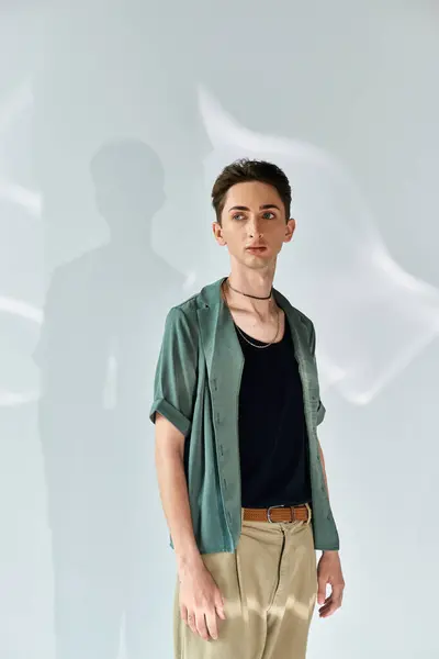 A young queer man strikes a confident pose in a stylish green shirt and tan pants against a white wall in a studio setting. — Stock Photo