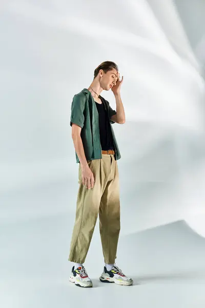 A young queer person in a tan shirt and khaki pants stands confidently against a plain white backdrop. — Stock Photo