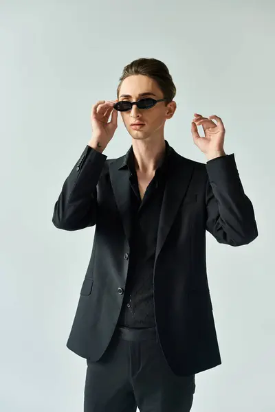 Uma jovem queer faz uma pose confiante em um terno preto e óculos de sol contra um fundo cinza. — Fotografia de Stock